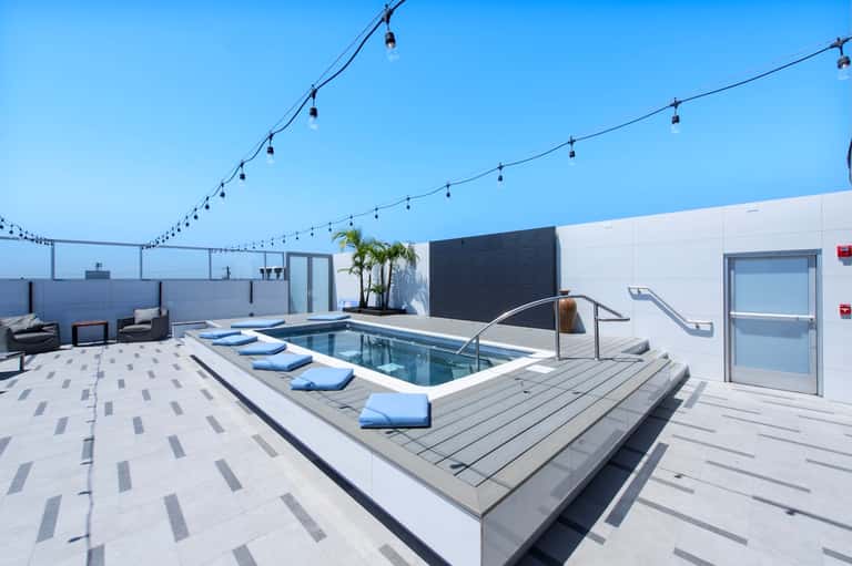 Modern rooftop pool deck with hot tub, blue loungers, string lights, and glass barriers