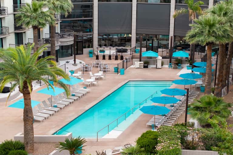 Resort pool surrounded by turquoise umbrellas, palm trees, loungers, and modern building facades