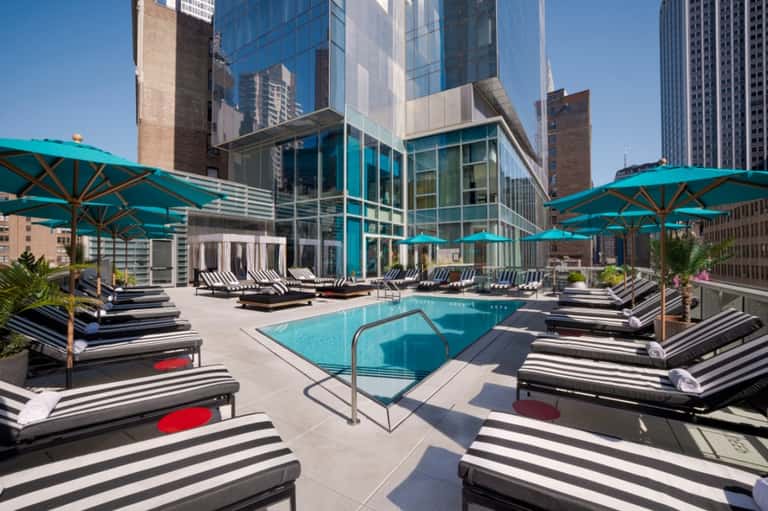 Rooftop pool with turquoise umbrellas, striped loungers, modern buildings, and city skyline views
