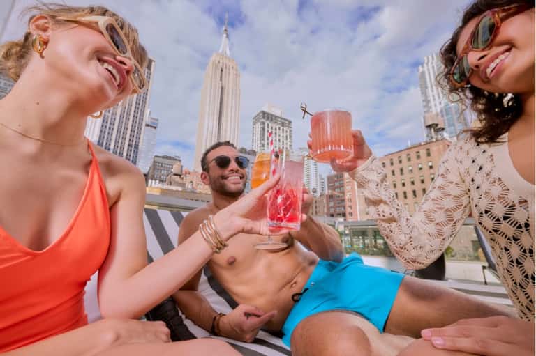Group of people on urban rooftop with Empire State Building visible in background, holding drinks and socializing