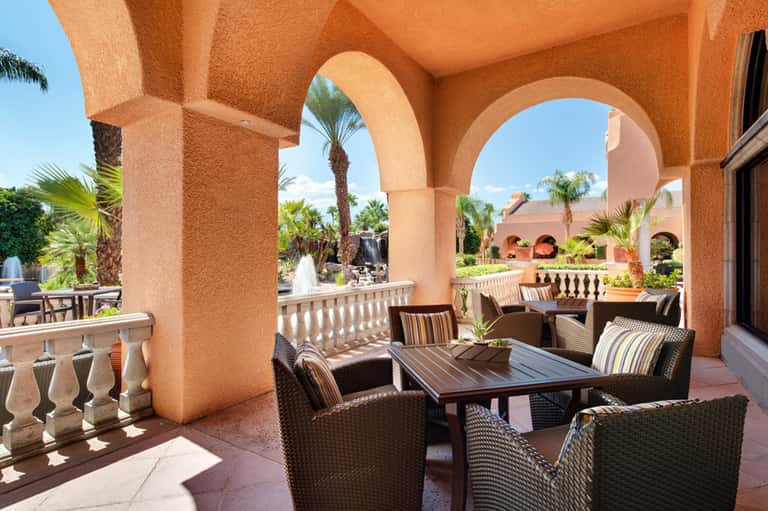 Terracotta-colored Spanish colonial architecture with arched covered patio, wicker seating, and tropical palm gardens