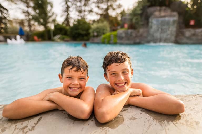 Two children smiling in resort pool with water slide and stone structure in background