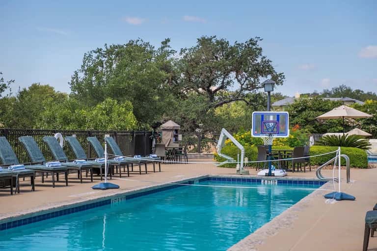 Resort pool with basketball hoop, loungers under blue umbrellas, and mature oak trees in background