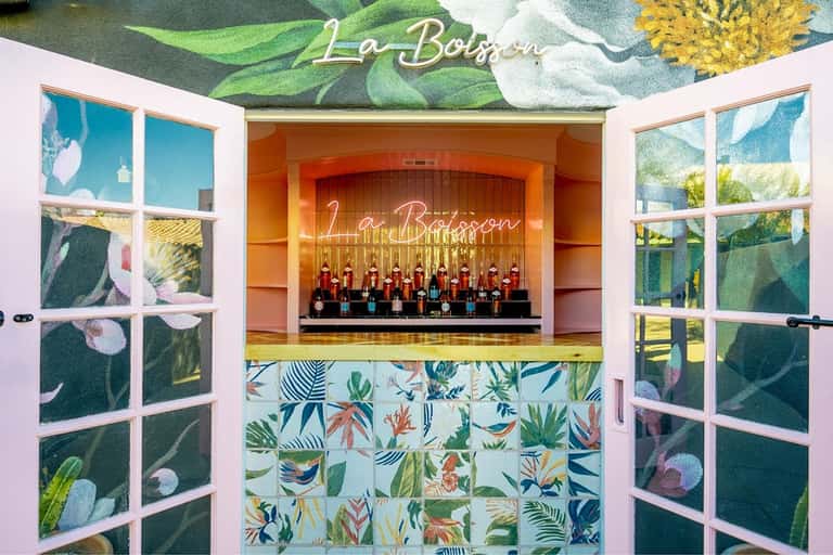 La Boisson bar with tropical botanical mural, pink doors, patterned tile counter, and bottle display