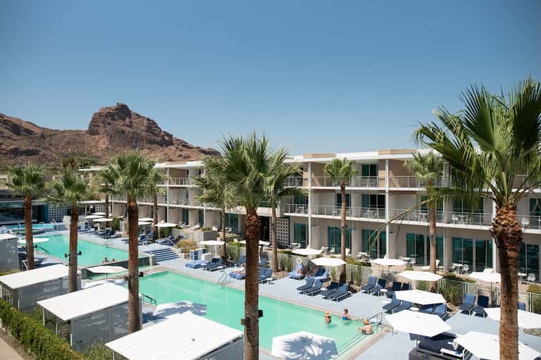 Resort aerial view with turquoise pools, white loungers, palm trees, and desert mountain backdrop