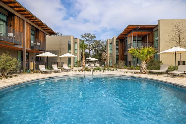 Resort courtyard with modern buildings, blue pool, white umbrellas, and palm trees