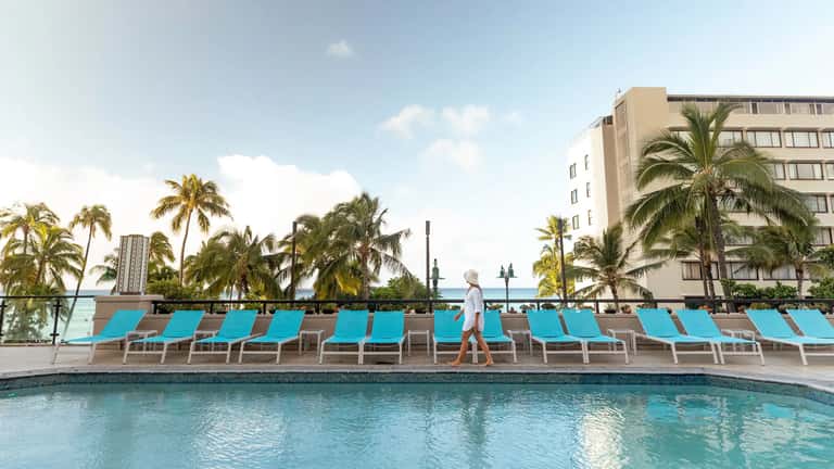 Resort pool with turquoise lounge chairs, palm trees, and ocean view in background