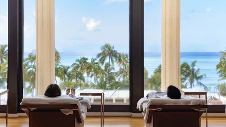 Spa treatment room with massage beds overlooking ocean and palm trees through large windows