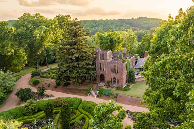 Aerial view of historic brick mansion surrounded by manicured formal gardens and lush forest landscape