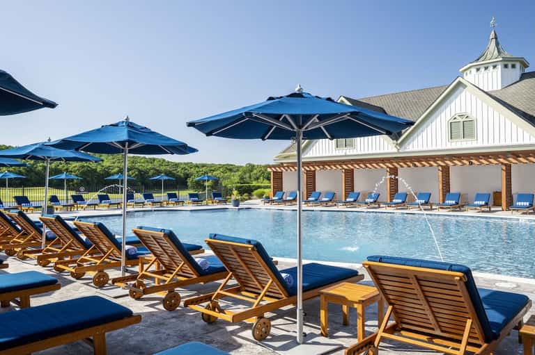 Resort pool with blue lounge chairs, umbrellas, white clubhouse, and manicured grounds