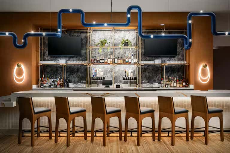 Modern bar with blue neon pipe design, wood paneling, multiple screens, and leather bar stools