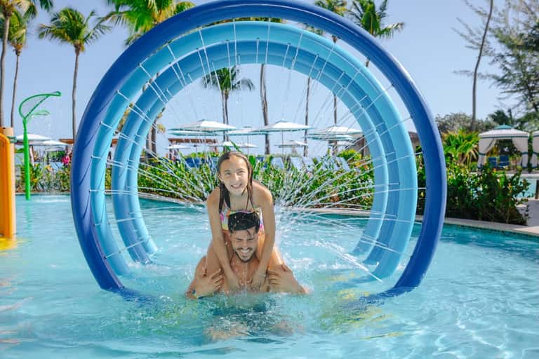 Father and daughter playing in pool through large blue tunnel toy with palm trees and umbrellas