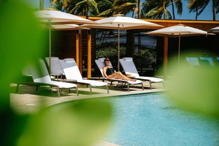 Modern poolside cabana with white lounge chairs, umbrella, wooden pergola, and palm trees framing the pool