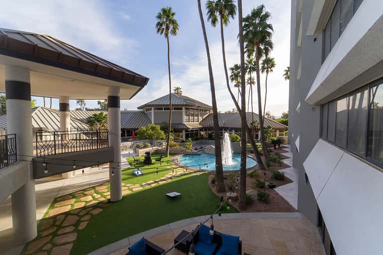 Resort courtyard with pool, manicured lawn, tall palm trees, covered pavilions, and modern architecture