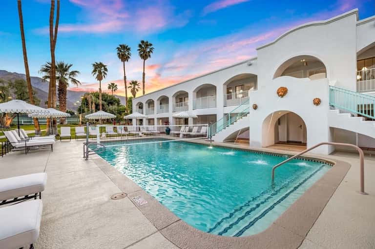 Resort pool with lounge chairs, umbrellas, palm trees, and white arched building at sunset