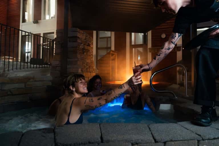 Guests enjoying illuminated hot spring pool at night with stone architecture and modern building