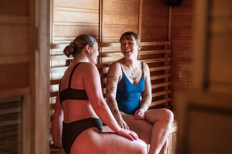 Two women relaxing in wooden sauna with warm lighting and wooden slat walls