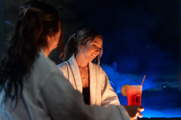 Two women in bathrobes enjoying drinks at nighttime with ambient lighting and ocean view in the background