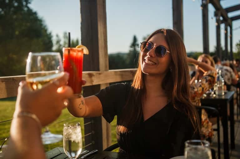 Woman holding colorful cocktails at outdoor bar with forest views and wooden pergola