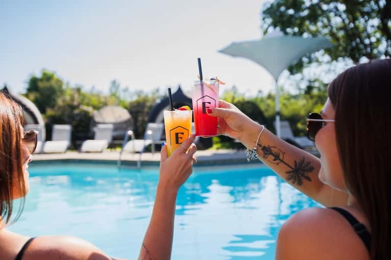 Two women toasting colorful cocktails poolside with resort lounge chairs visible in background