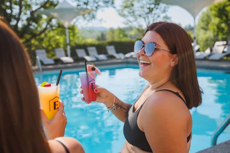 Woman in sunglasses holding colorful cocktails by the pool with lounge chairs and trees visible