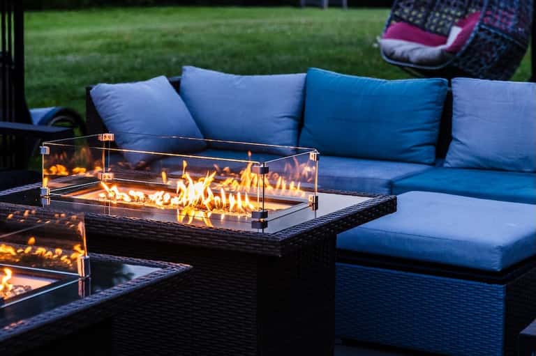 Outdoor patio with blue cushioned seating, fireplace table, and manicured lawn at dusk