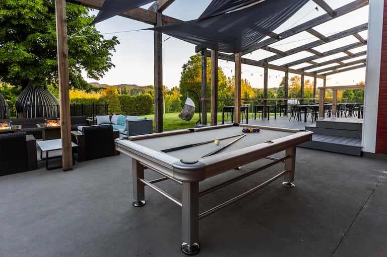 Modern covered patio with pool table, pergola shade structure, outdoor seating, and views of forested landscape.