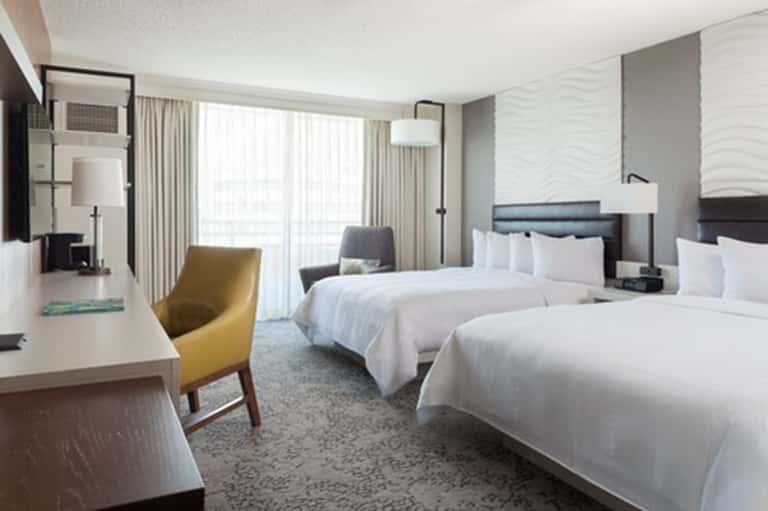 Modern hotel room with two beds, seating area, large windows, and neutral tones