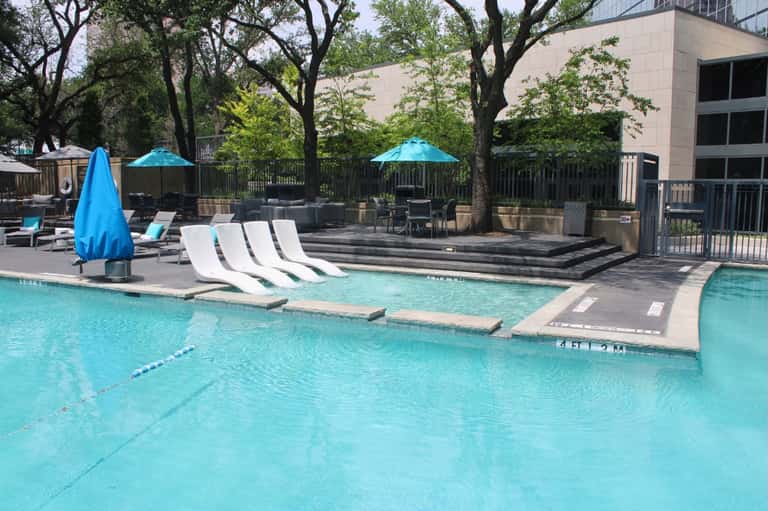 Resort pool with turquoise water, white lounge chairs, blue umbrellas, mature trees, and modern building