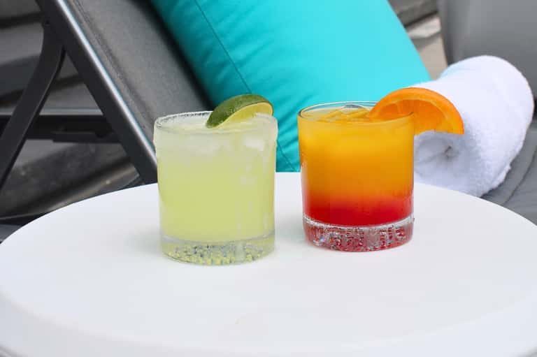 Two colorful cocktails with lime and orange garnish on white table by turquoise lounge chair
