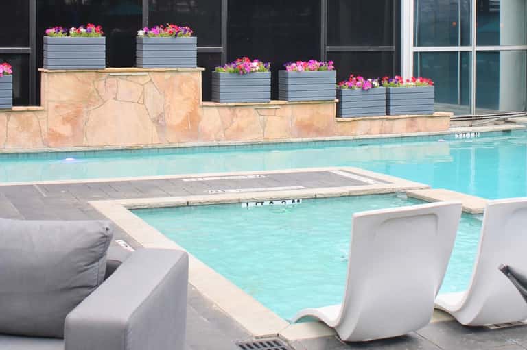 Resort pool with heated spa, pink flowers in planters, and lounge chairs against glass building