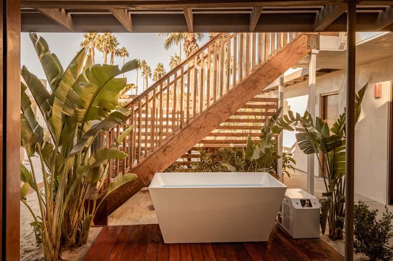 Outdoor bathroom with freestanding white bathtub, wooden stairs, and tropical plants on beach