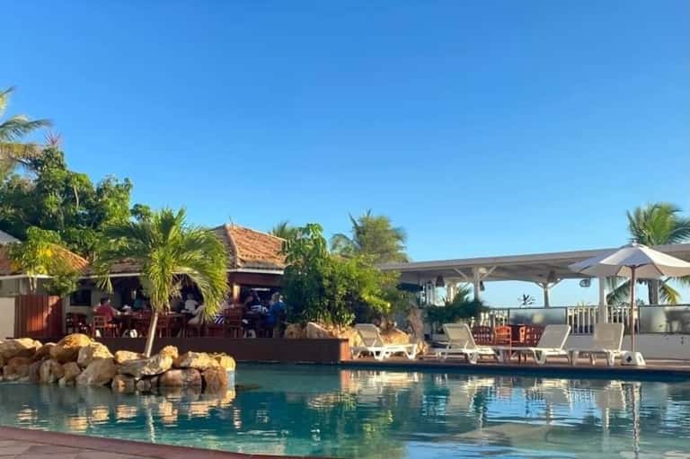 Tropical resort pool area with tiki bar, wooden lounge chairs, palm trees, and ocean view