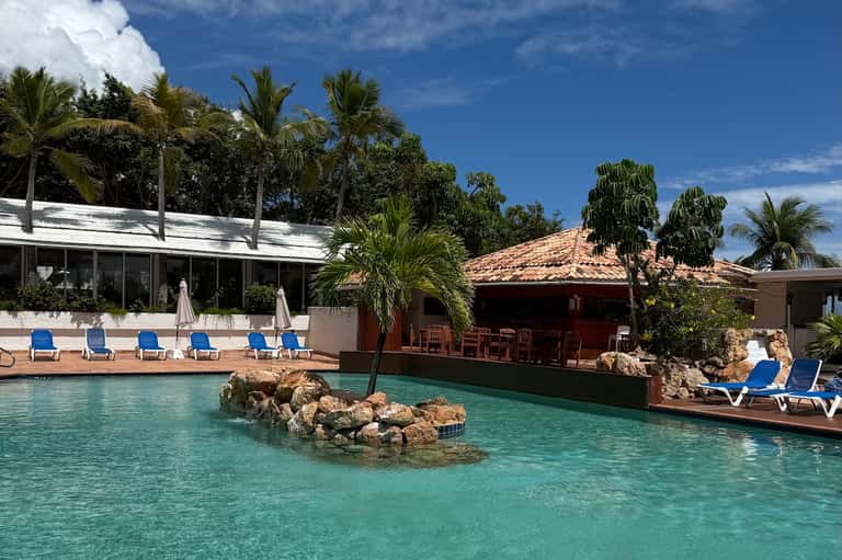 Resort pool with turquoise water, rocky islands, palm trees, and beachfront buildings