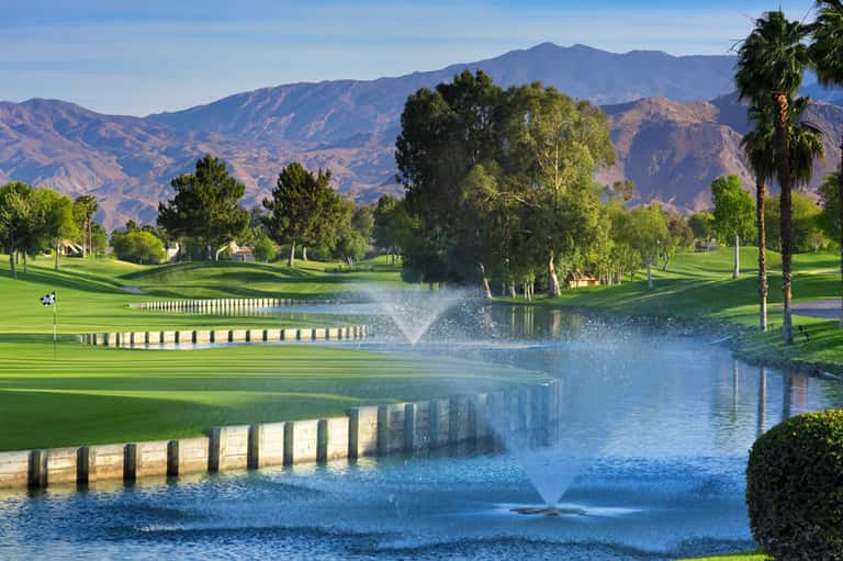 Championship golf course with manicured fairways, water hazard, fountain, and mountains in background