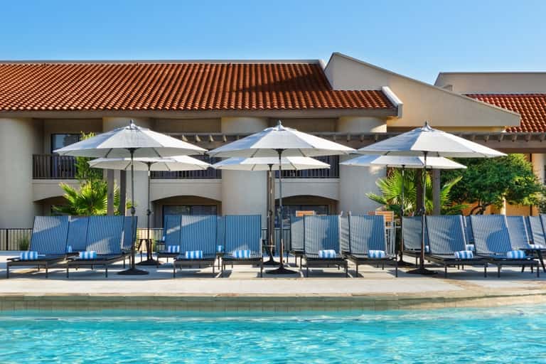 Resort poolside with white umbrellas, blue lounge chairs, clay tile roof building, and palm trees