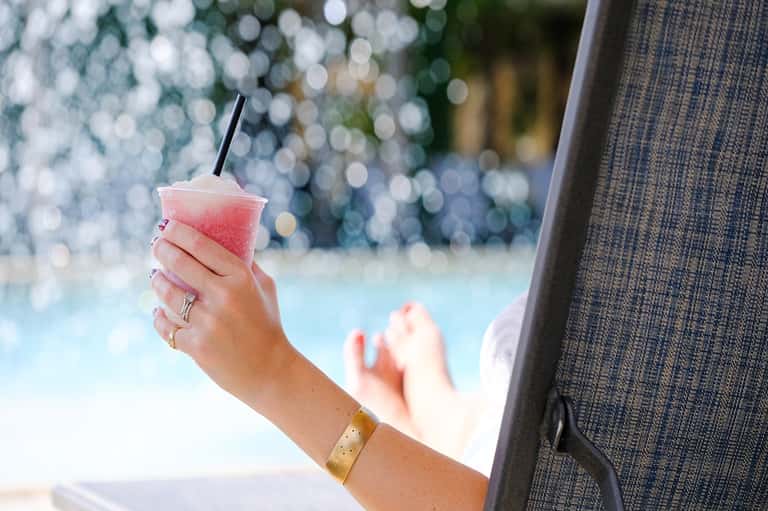 Woman's hand holding pink cocktail by pool with sparkling water in background