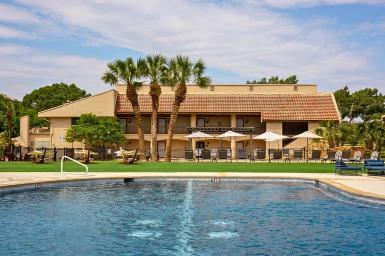 Resort pool with palm trees, beige building, and lounge chairs on green lawn