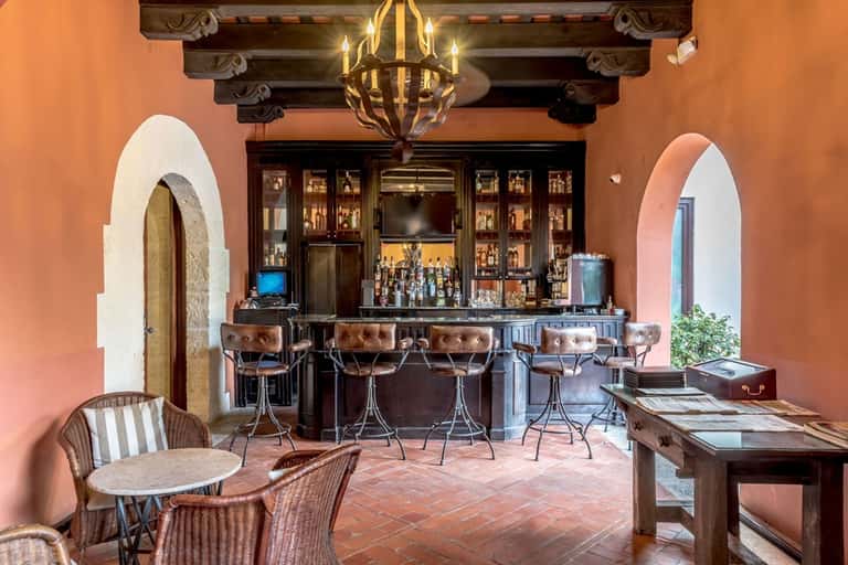 Upscale bar lounge with coral walls, dark bar counter, leather stools, and rustic wooden furnishings.