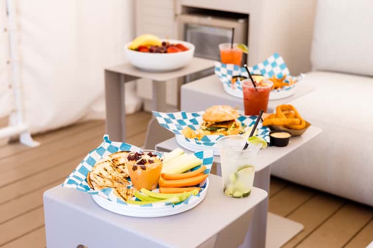 Outdoor dining setup with white plates, blue patterned napkins, burgers, beverages, and fresh fruit