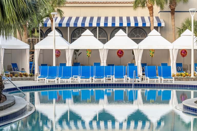 Resort pool deck with white cabanas, blue lounge chairs, palm trees, and striped awning