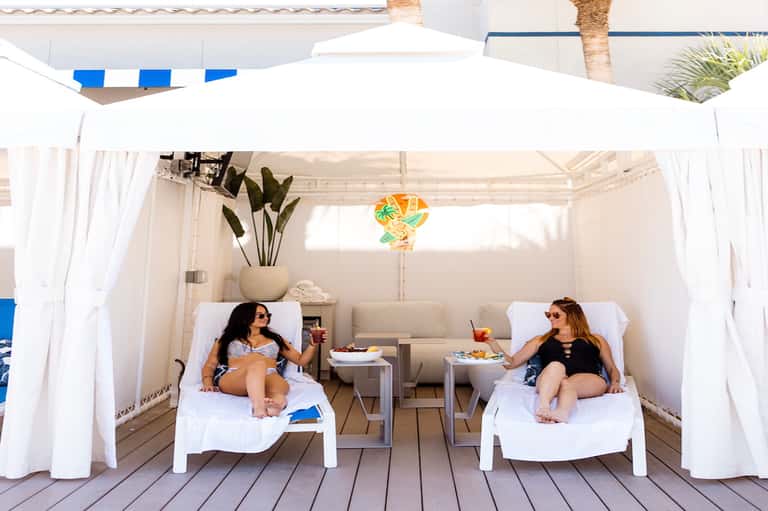 Two women lounging in white cabana beds under flowing white curtains and palm umbrella