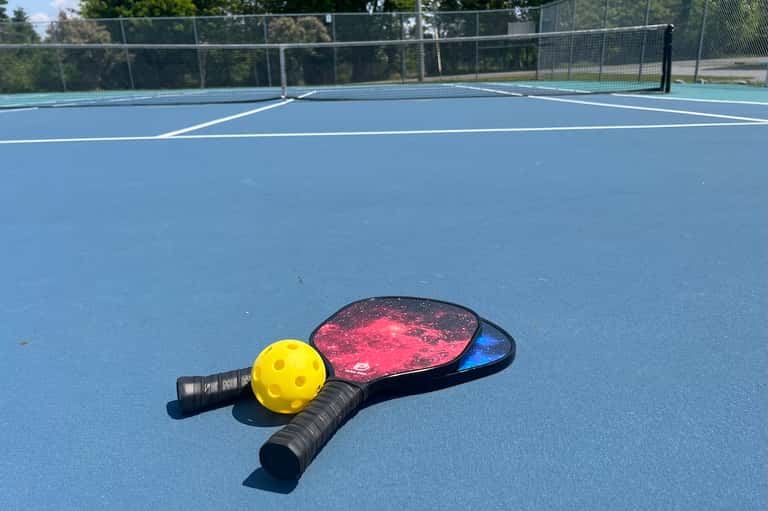 Pickleball paddle and yellow ball on blue court with net and fencing in outdoor recreation area