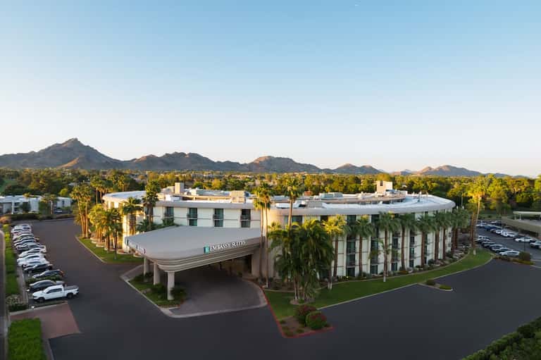 Modern Embassy Suites hotel with white facade and palm trees, mountains visible in distance