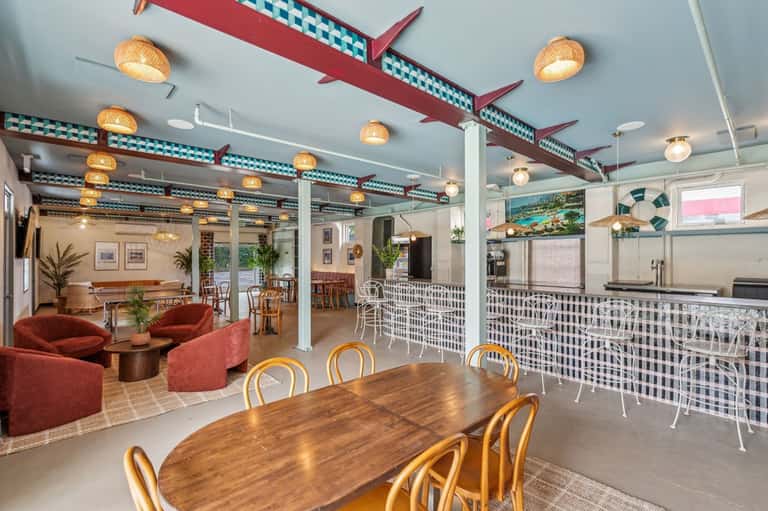 Bright resort club room with teal ceiling beams, gold pendant lights, red seating areas, white railings, and dining tables