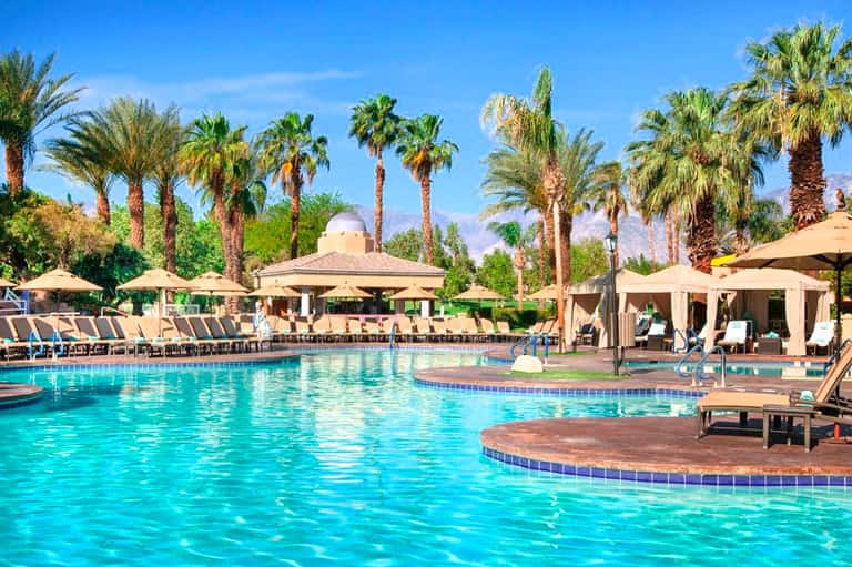 Resort pool with crystal-clear water, palm trees, cabanas, and loungers under umbrellas on a sunny day