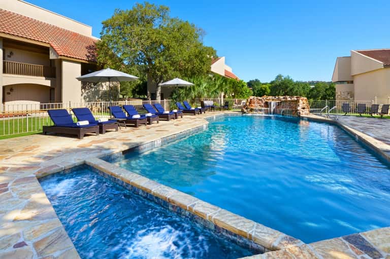 Resort pool with spa, blue lounge chairs, umbrellas, waterfall feature, and surrounding residential buildings