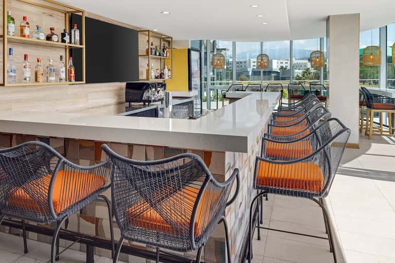 Rooftop bar with city skyline views, modern wicker bar chairs with orange cushions, wooden shelving, and floor-to-ceiling windows