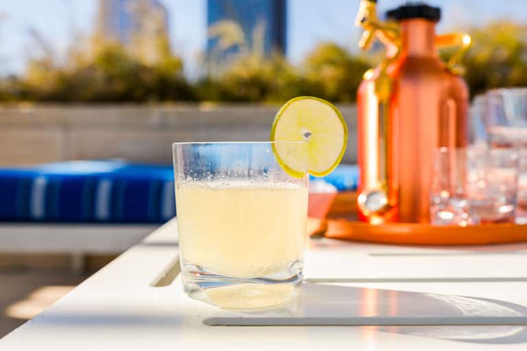 Pale yellow cocktail with lime garnish on white surface near resort pool with blurred background