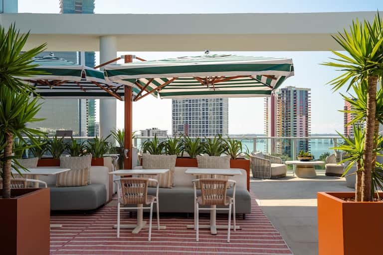 Rooftop lounge area with green striped umbrellas, seating, tropical plants, and city/waterfront views